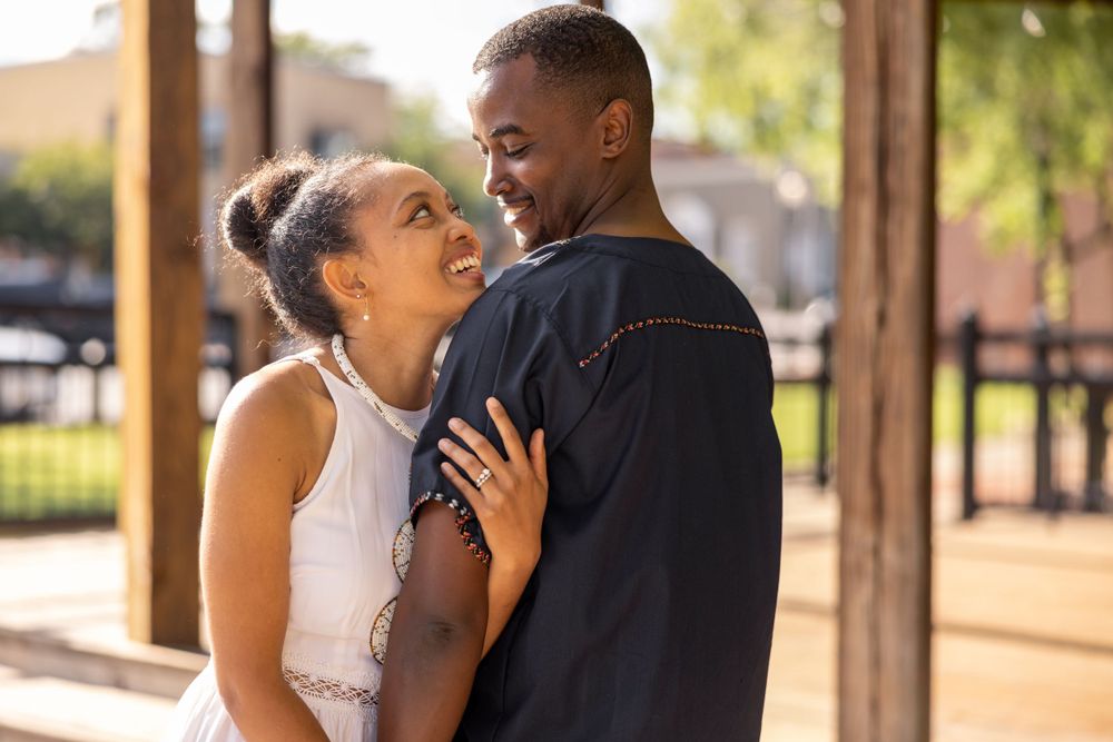 A couple shares a loving embrace in warm evening sunlight under a wooden structure.