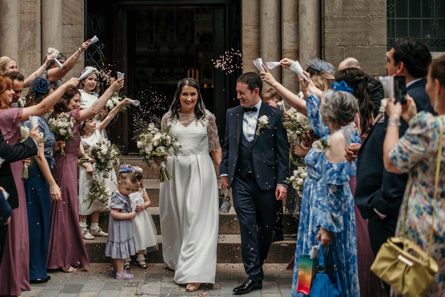 Guests line up outside a grand building to shower newlyweds with confetti.