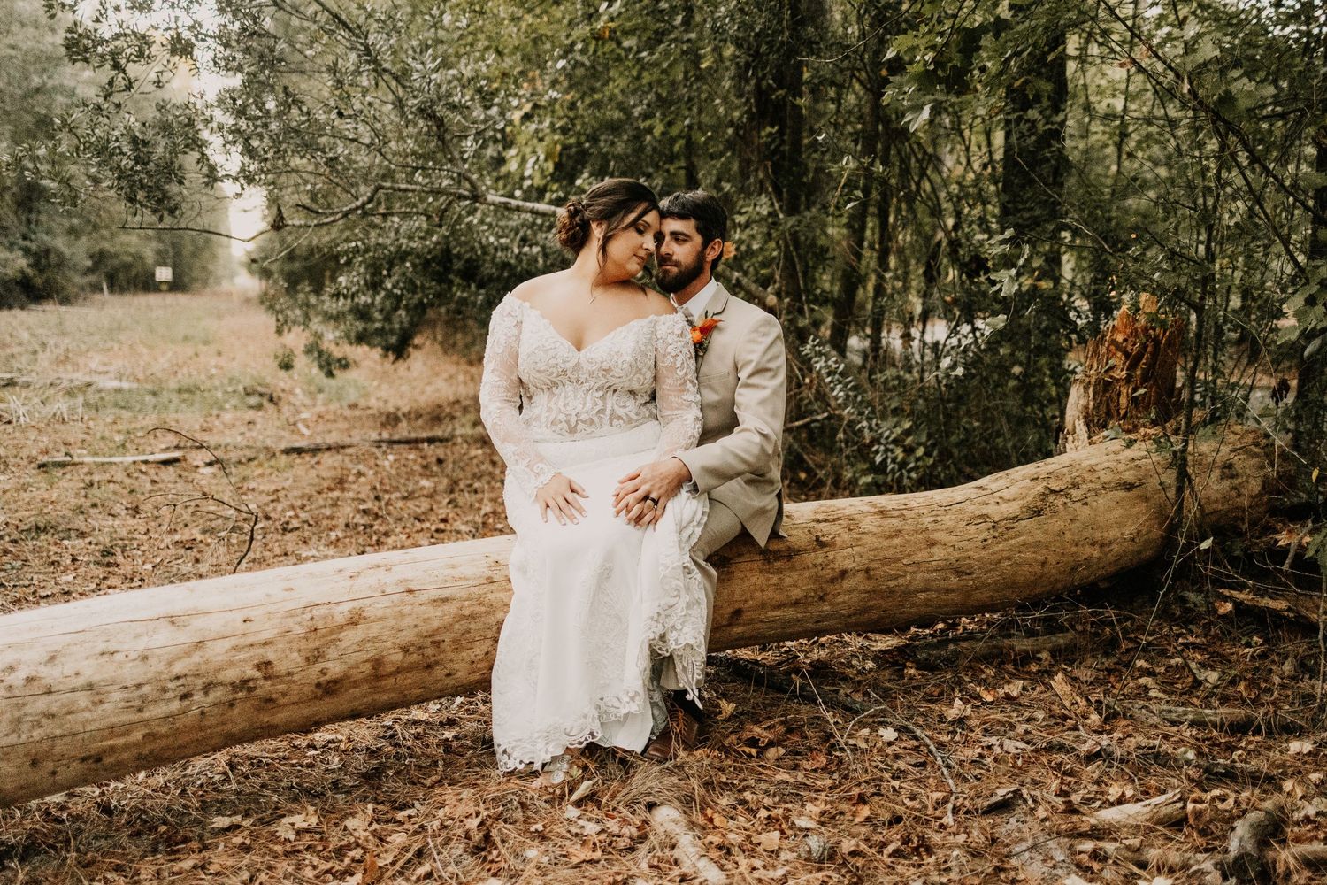 Couple sits together on fallen log in forest wearing romantic wedding attire.