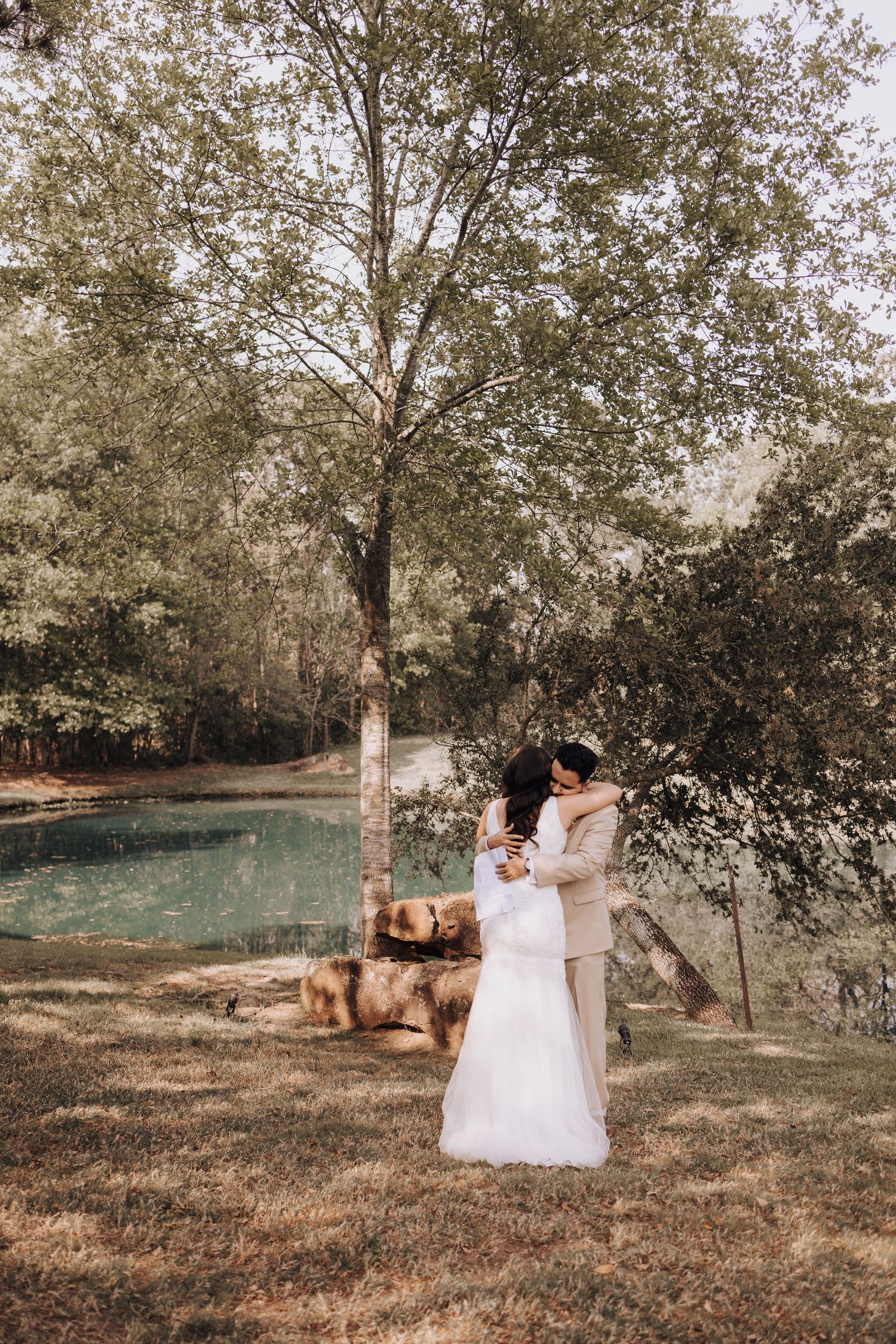 Romantic wedding moment captured under tall trees beside a turquoise pond.