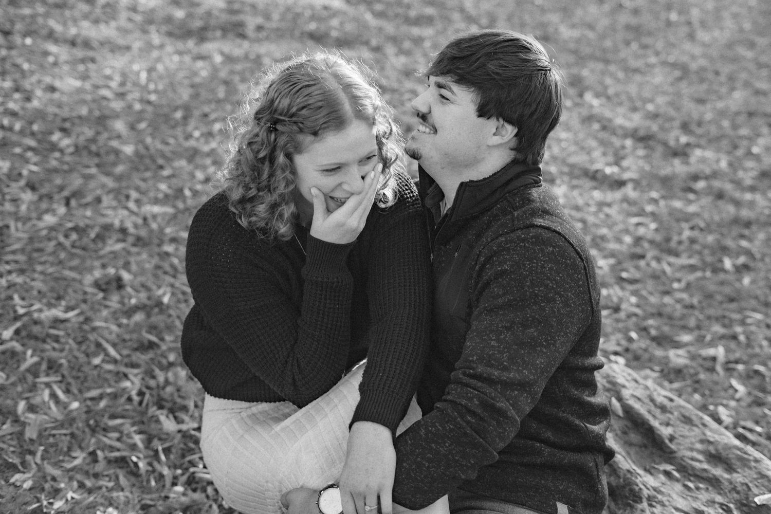 a woman covering her mouth and sitting on a mans lap on the grass while they both laugh