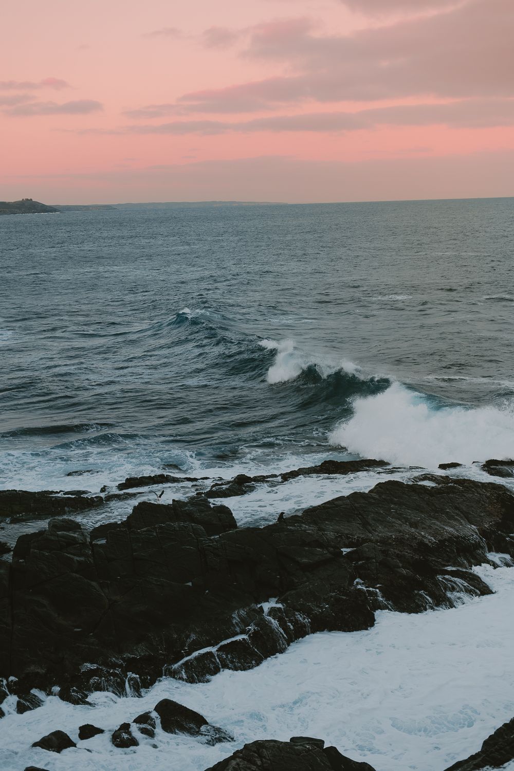 Coastal seascape at sunset with waves crashing against rocky shore under pink sky.