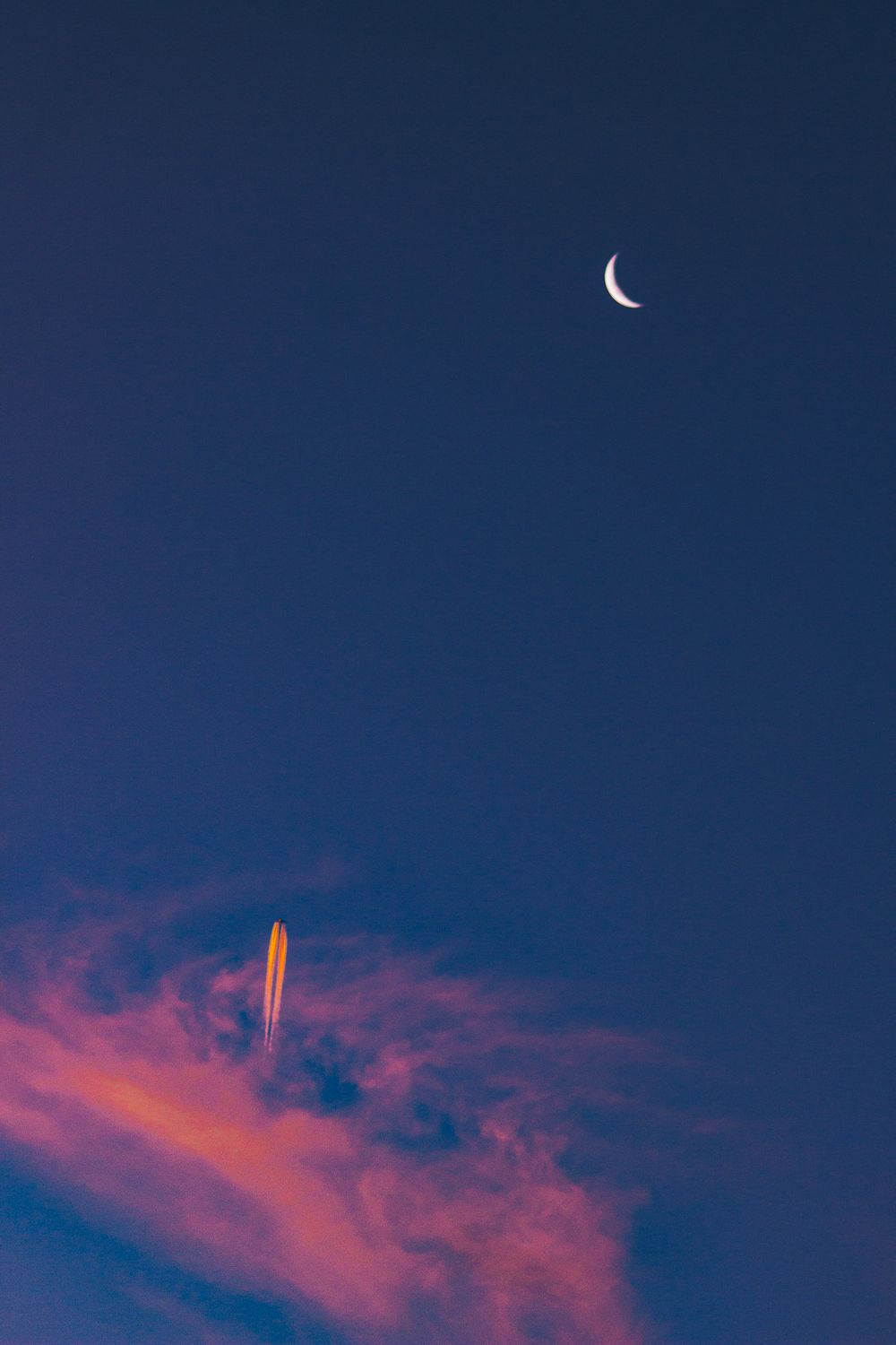 Crescent moon in a deep blue sky with pink wispy clouds at dusk.