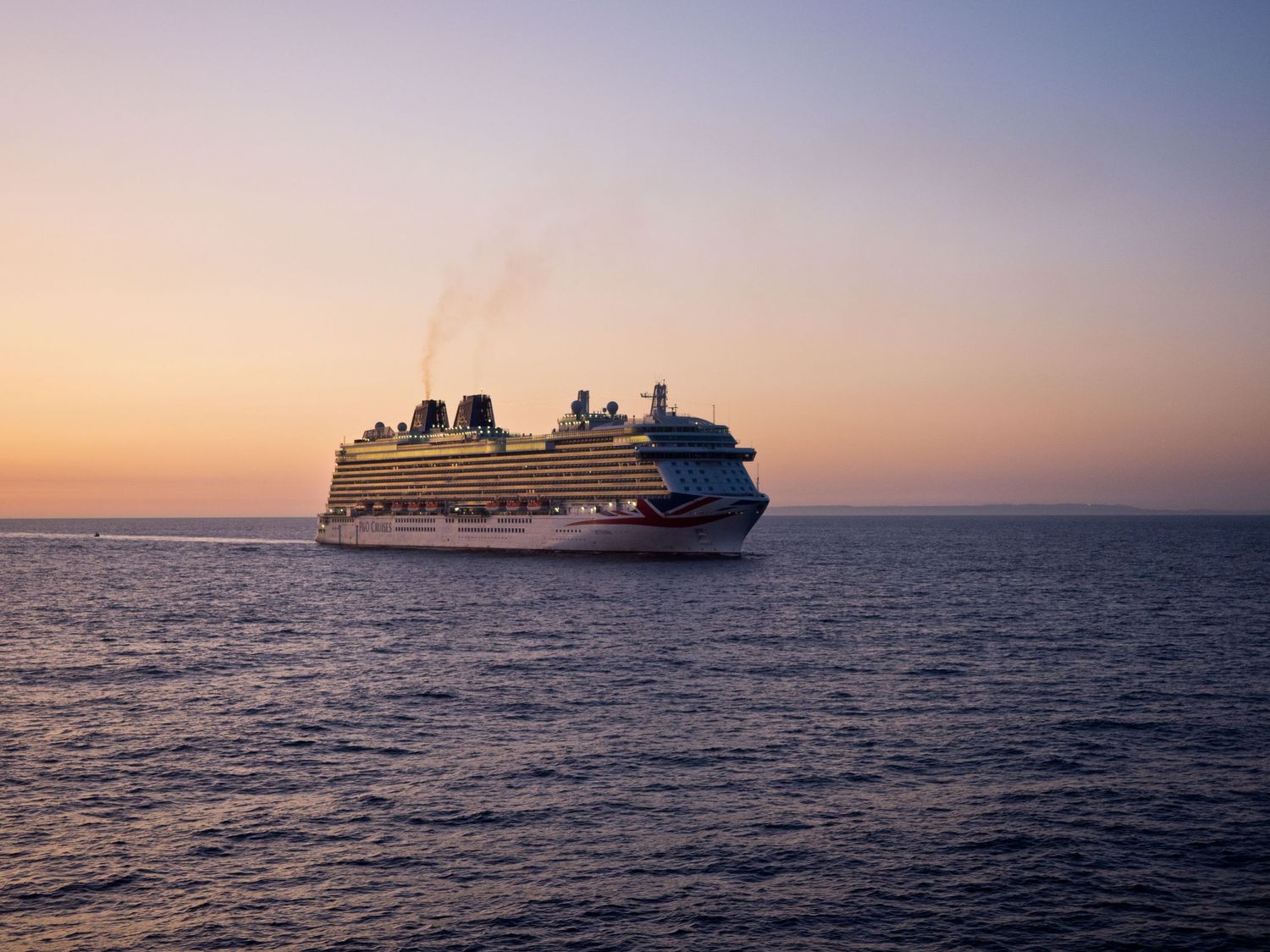 Large cruise ship silhouetted against a sunset sky over calm ocean waters.