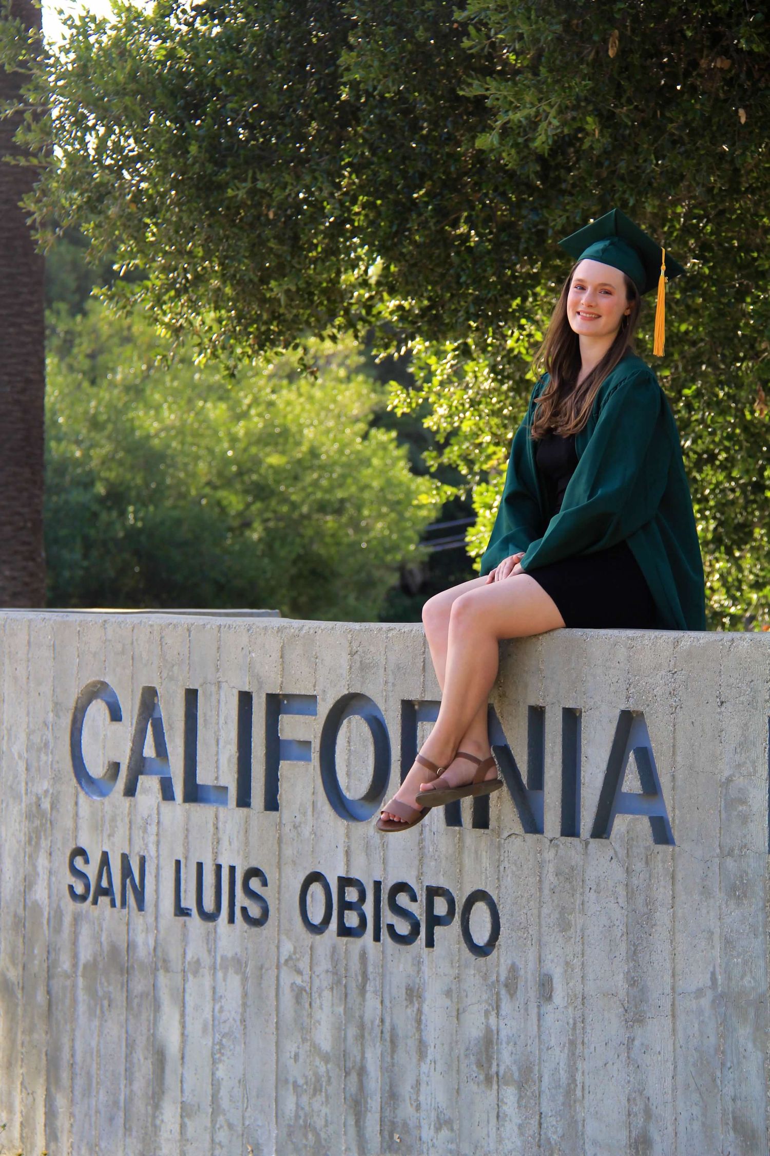 Graduate in green cap and gown sits on California San Luis Obispo campus sign.