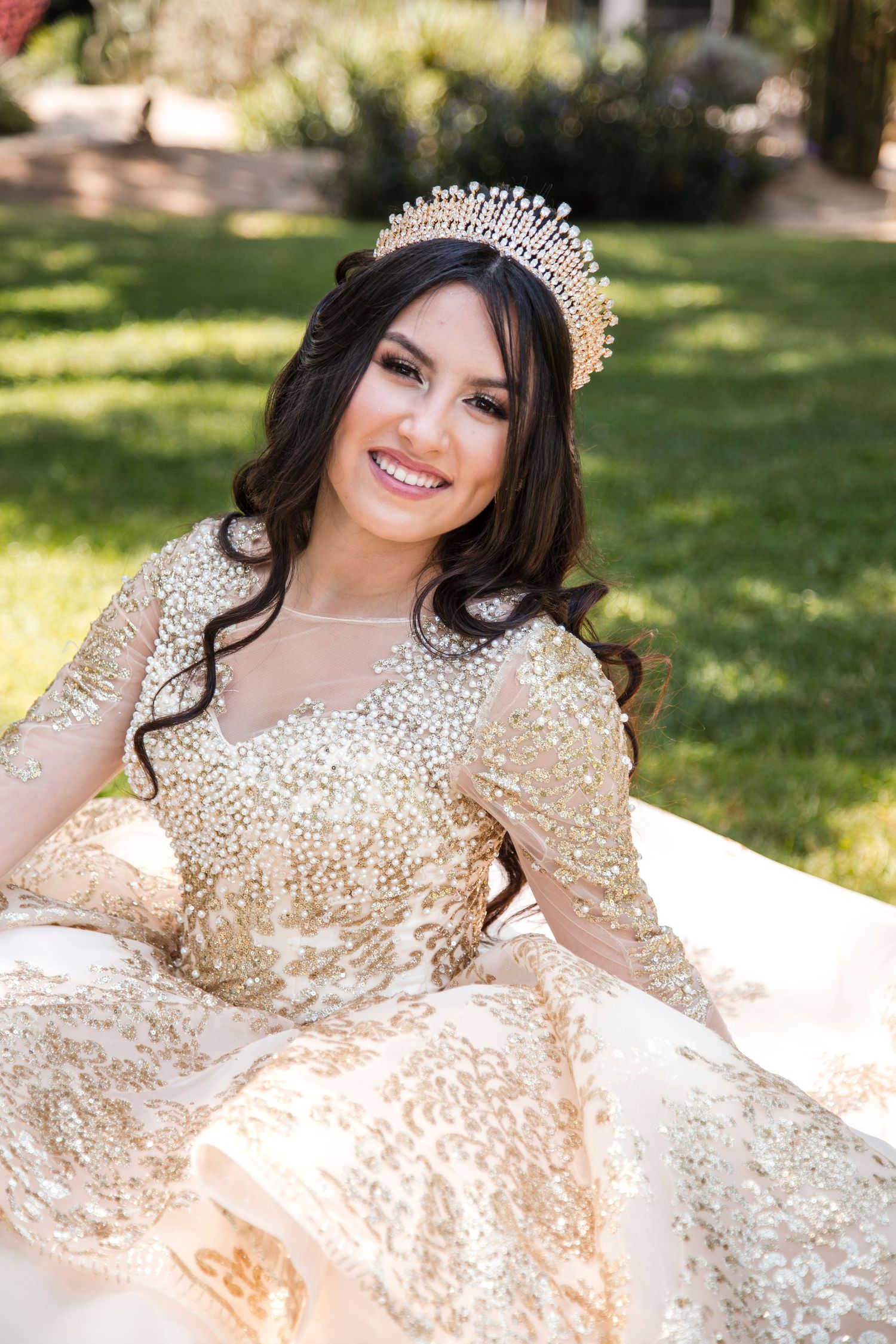 Person wearing a sparkling gold gown and tiara posing on grass in natural sunlight.