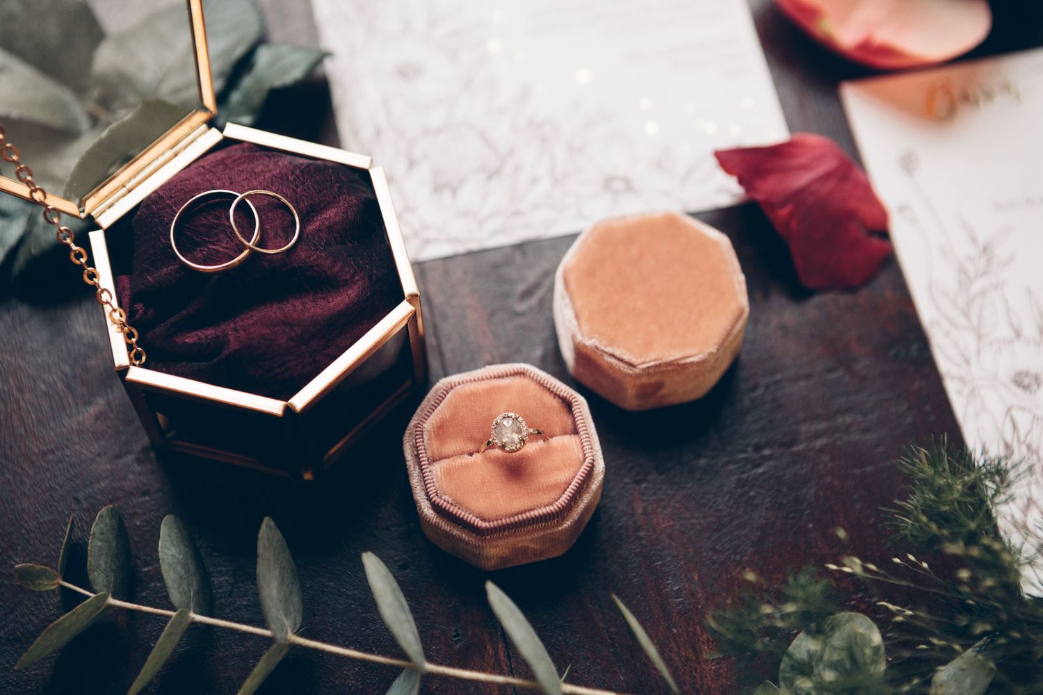 Wedding rings and engagement ring displayed in velvet hexagonal jewelry boxes on dark wooden surface with greenery.