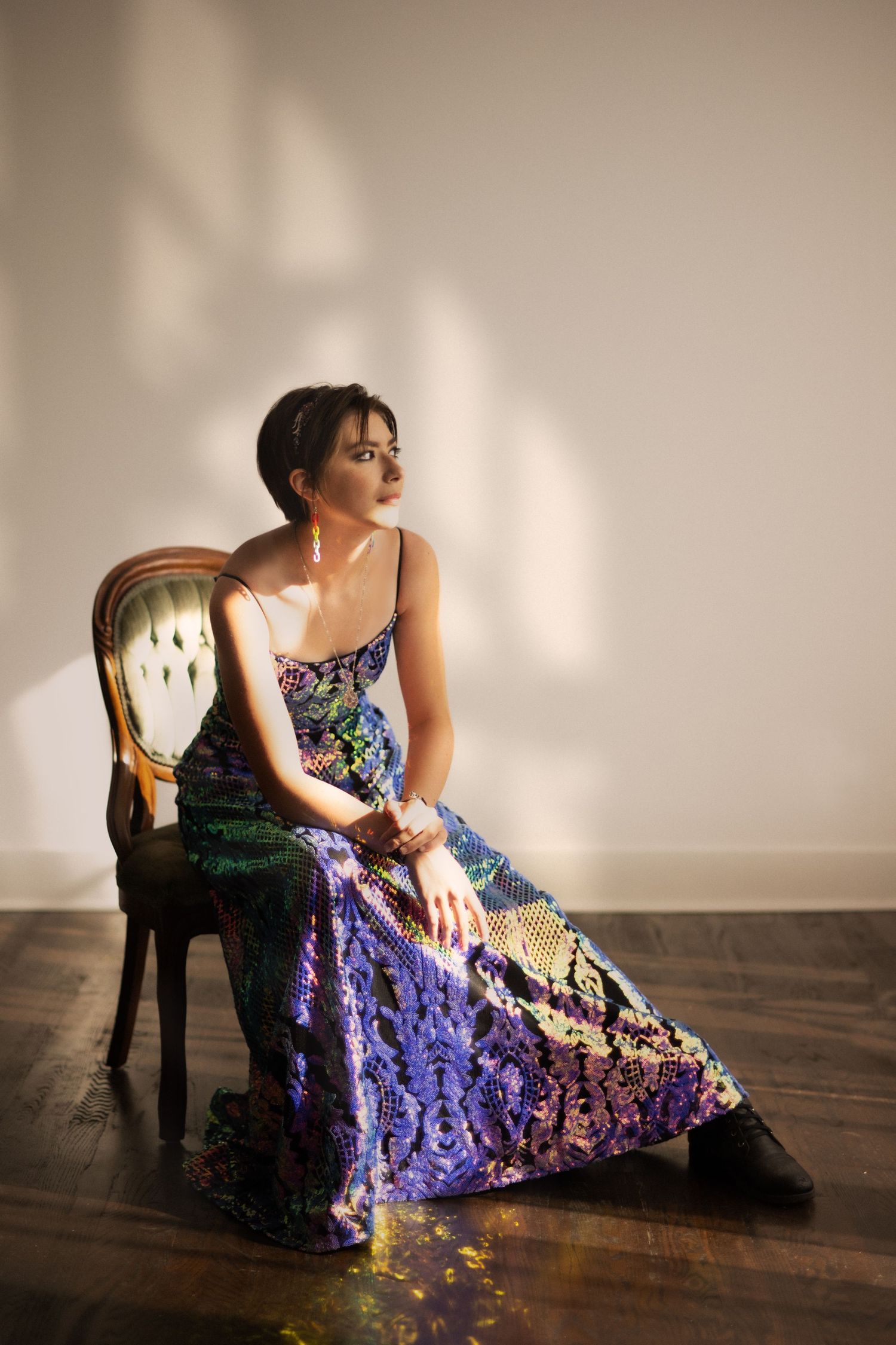 A model in a vibrant blue and purple sequined gown poses elegantly on an antique chair against a minimalist backdrop.
