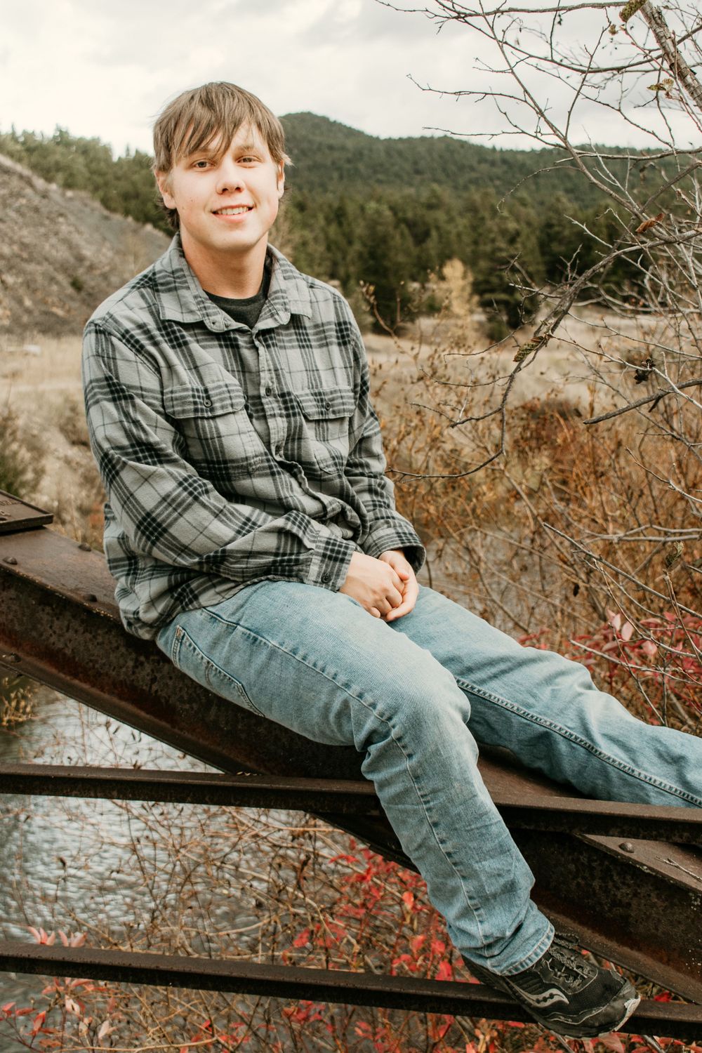Person in gray plaid shirt sits casually on wooden railing with mountain scenery behind.