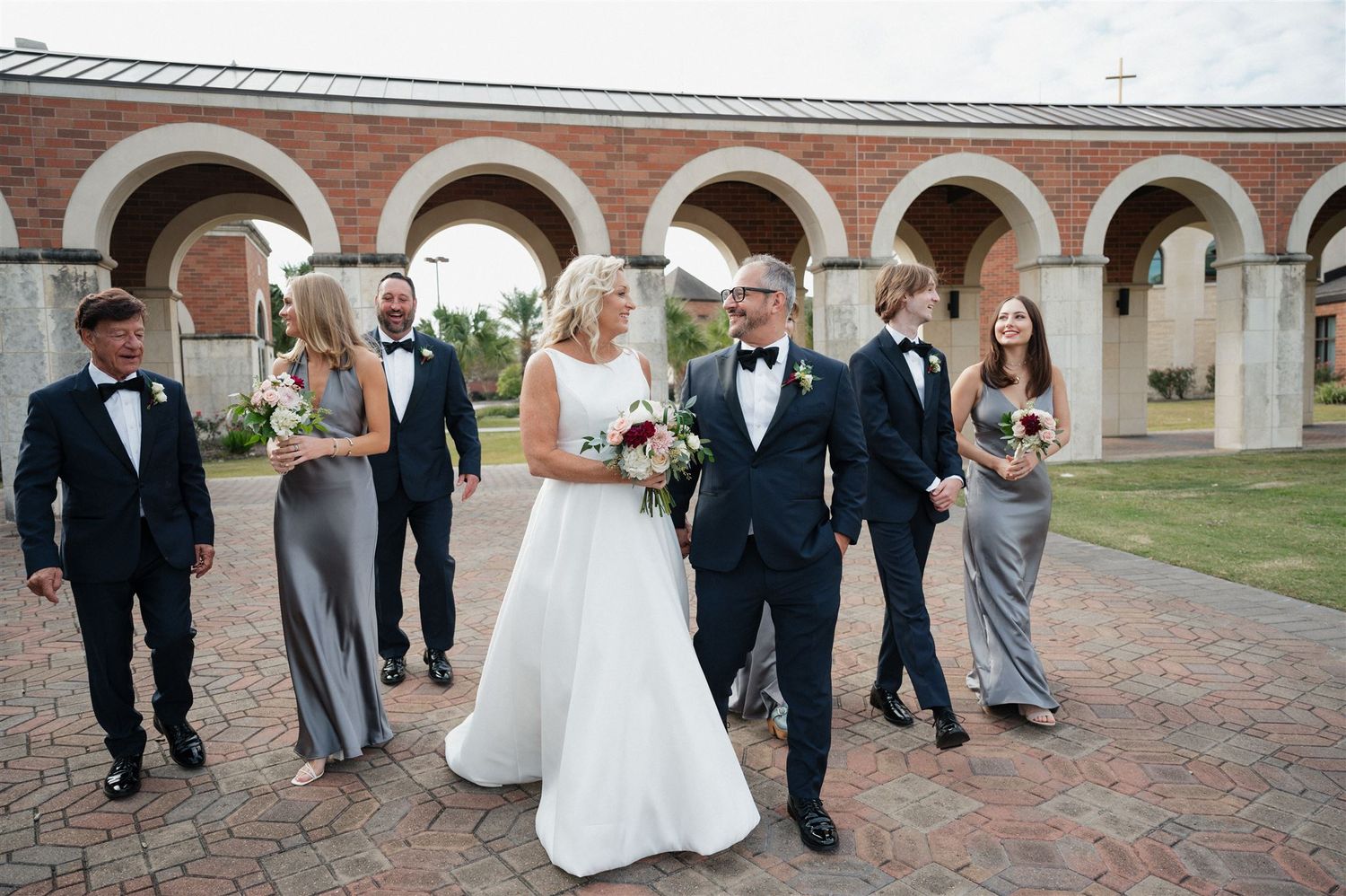 Wedding group walks casually along a covered walkway with brick arches.