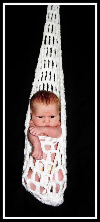 Newborn baby nestled in white macrame hanging basket against black background.