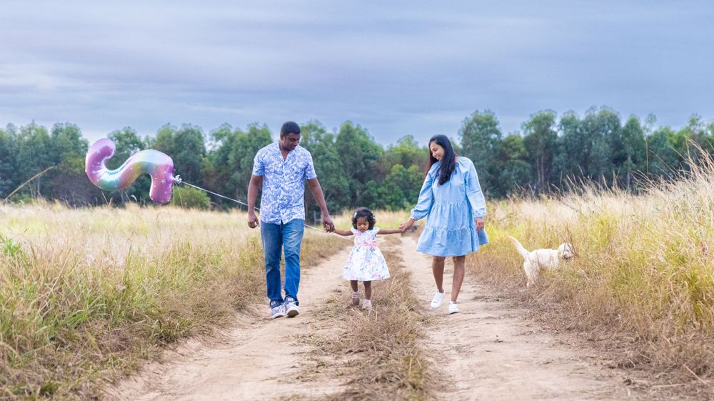 Family strolling down a rural path with balloons and their white dog in golden light.