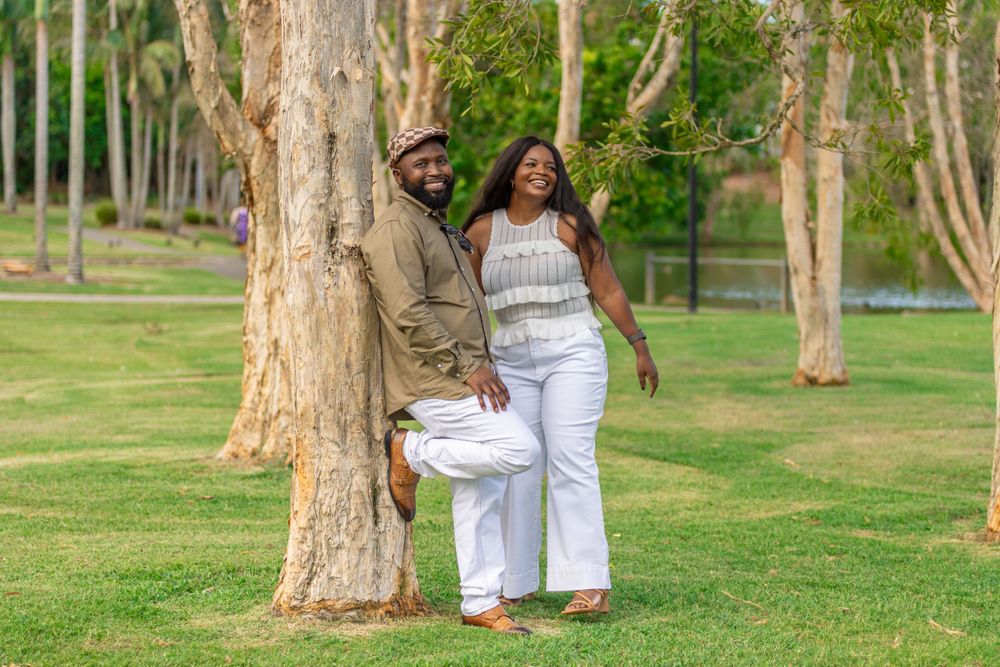 A couple in casual clothing poses playfully by a tree in a park setting with green grass.