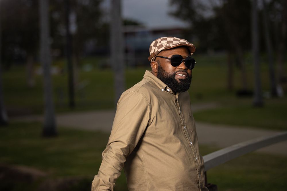 Person in a tan jacket and patterned hat poses outdoors at sunset.