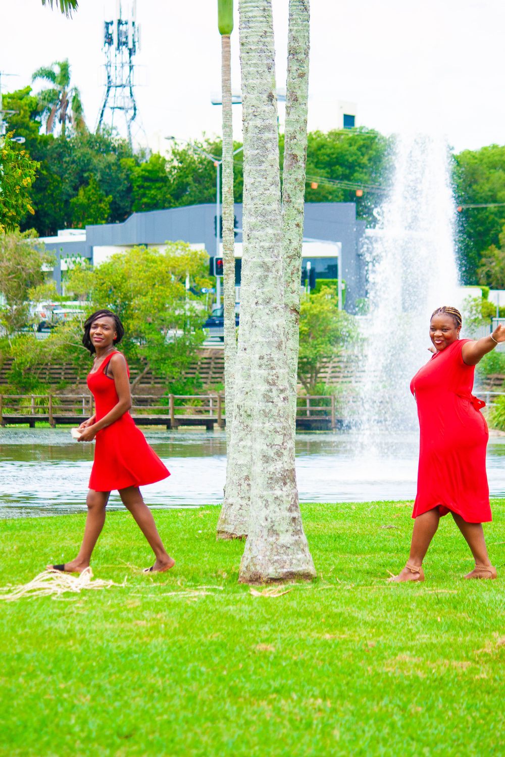 Two people in red dresses strike playful poses by palm trees and fountain.
