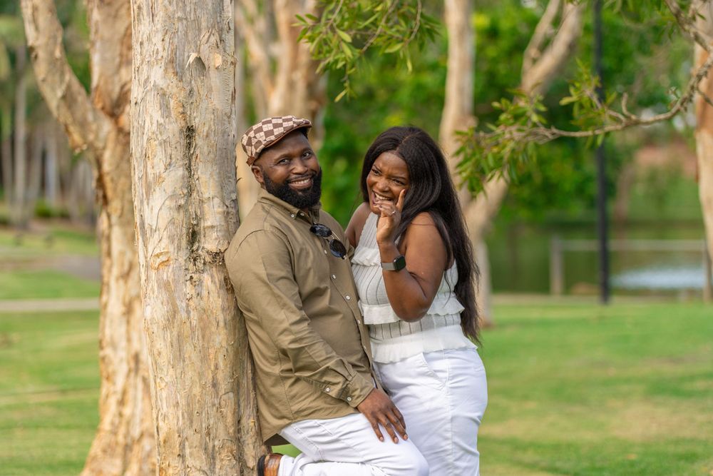A happy couple laughs together while posing against a tree trunk in an outdoor setting.