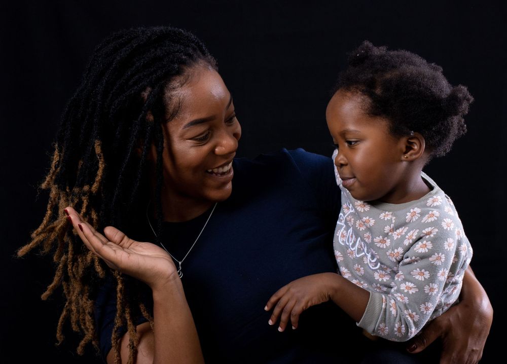 Person with dreadlocks smiling while holding small child in patterned outfit.