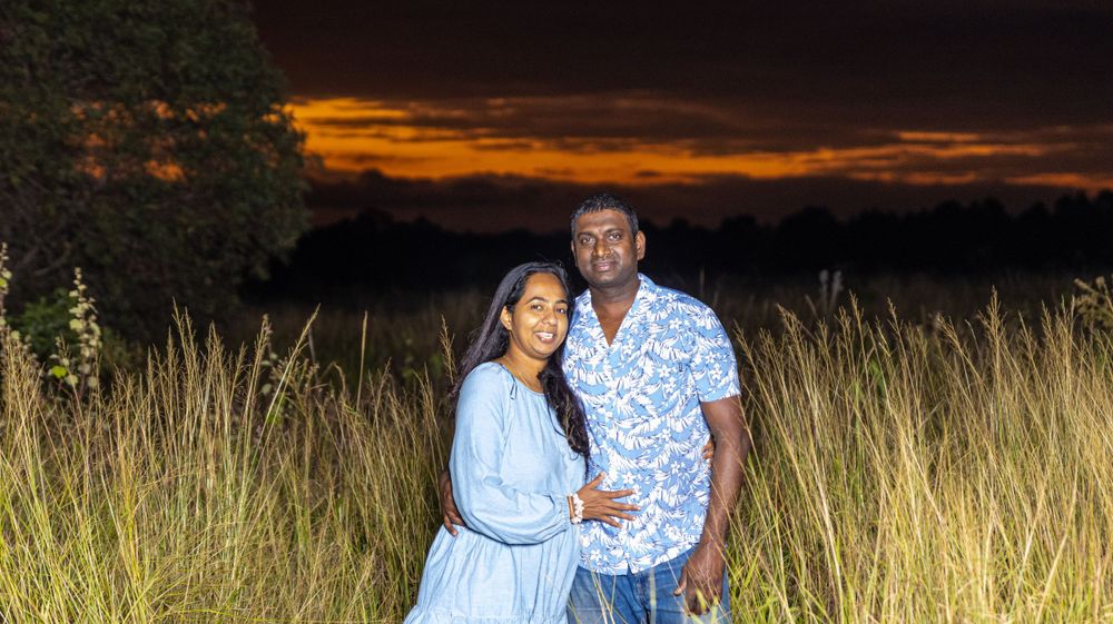 A couple poses in tall grass against a dramatic orange sunset sky in a romantic outdoor setting.