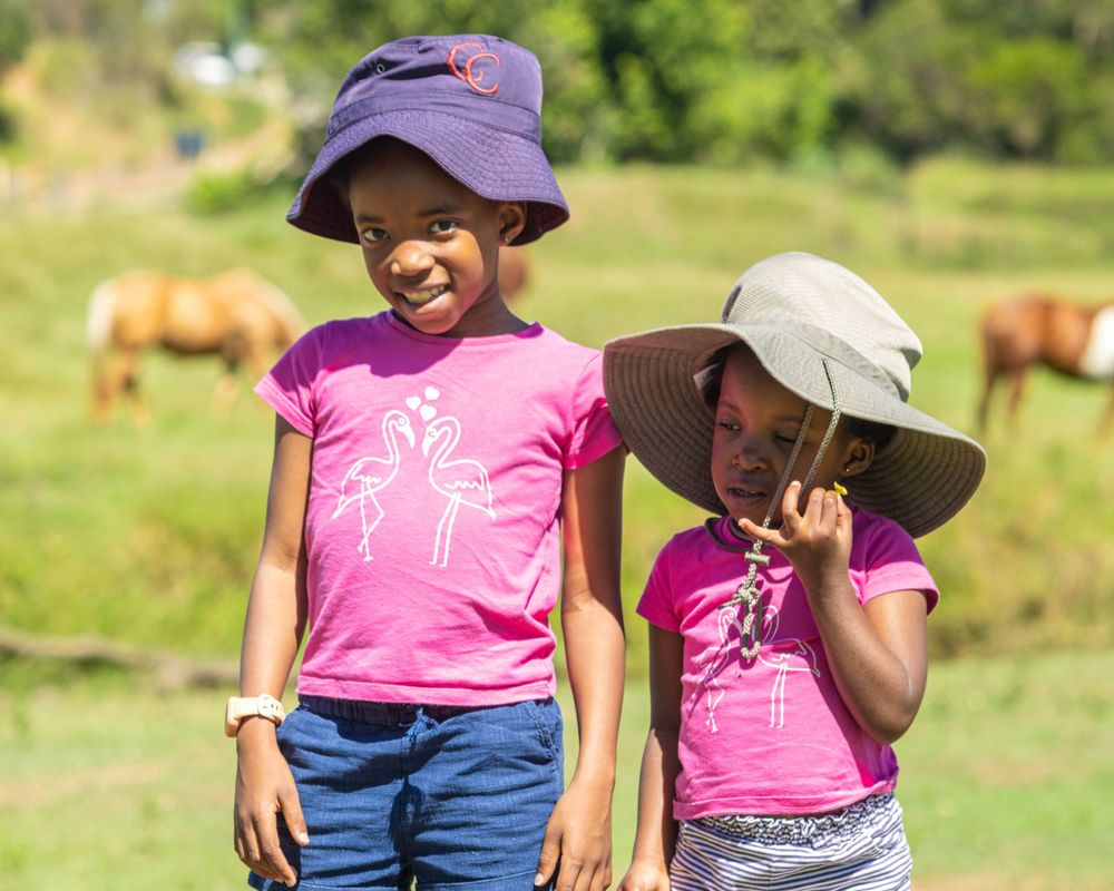 Two children wearing purple hats and pink shirts smile in sunny field with cattle.