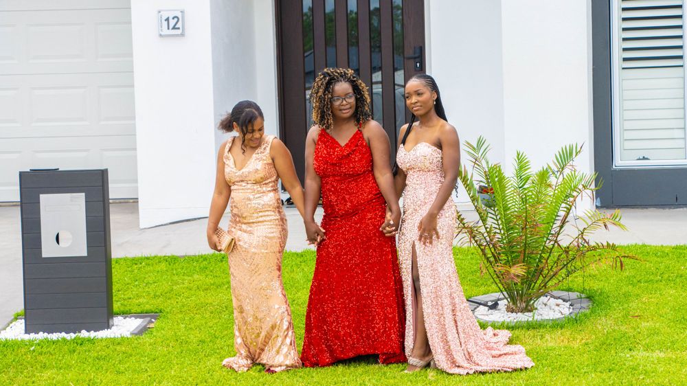 Three people wearing elegant evening gowns pose on a manicured lawn outside a modern home.