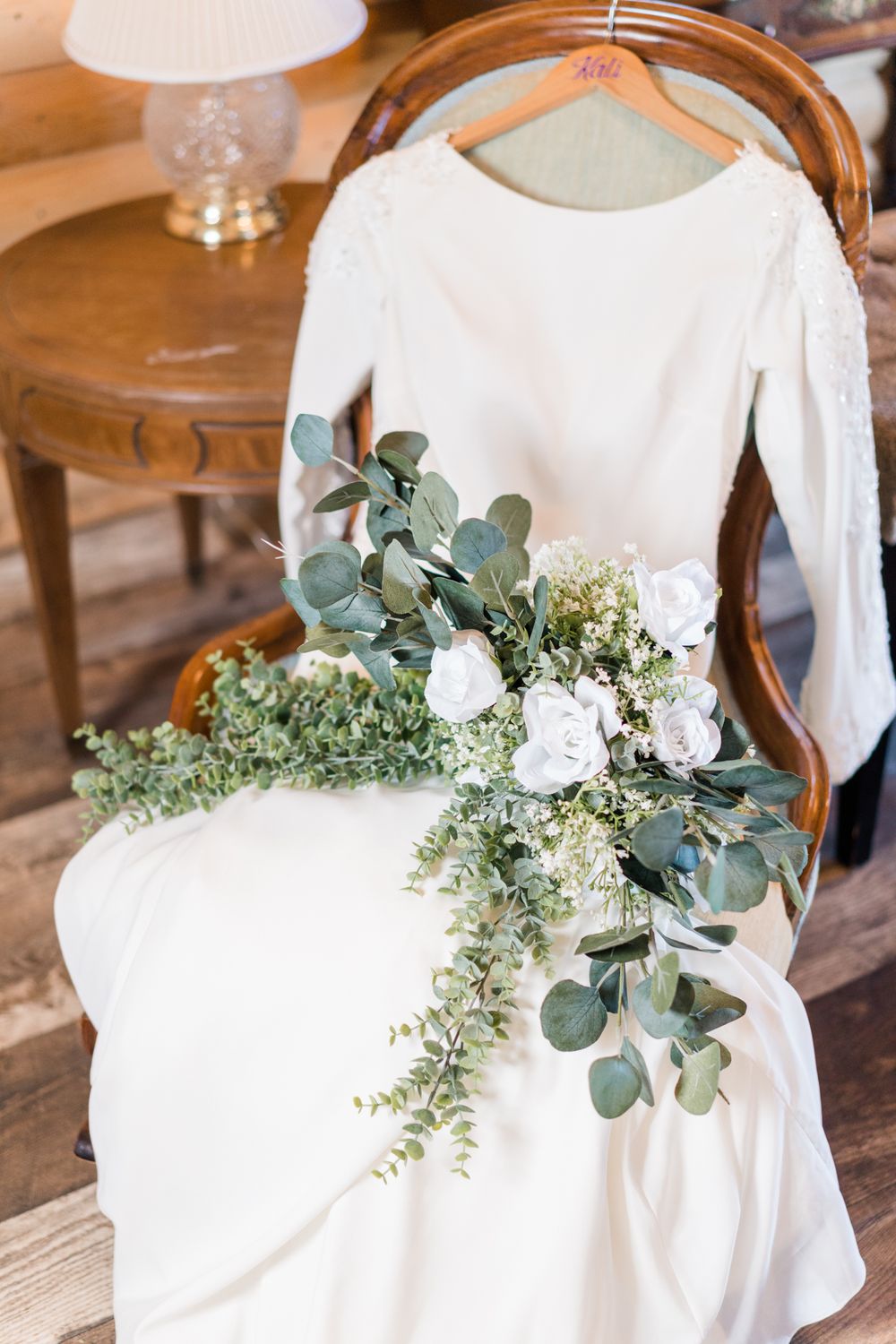 White wedding dress on hanger with eucalyptus and floral bouquet.