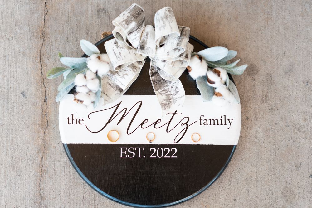Round wooden family name sign decorated with white flowers and greenery reading 'The Meetz Family Est. 2022'.