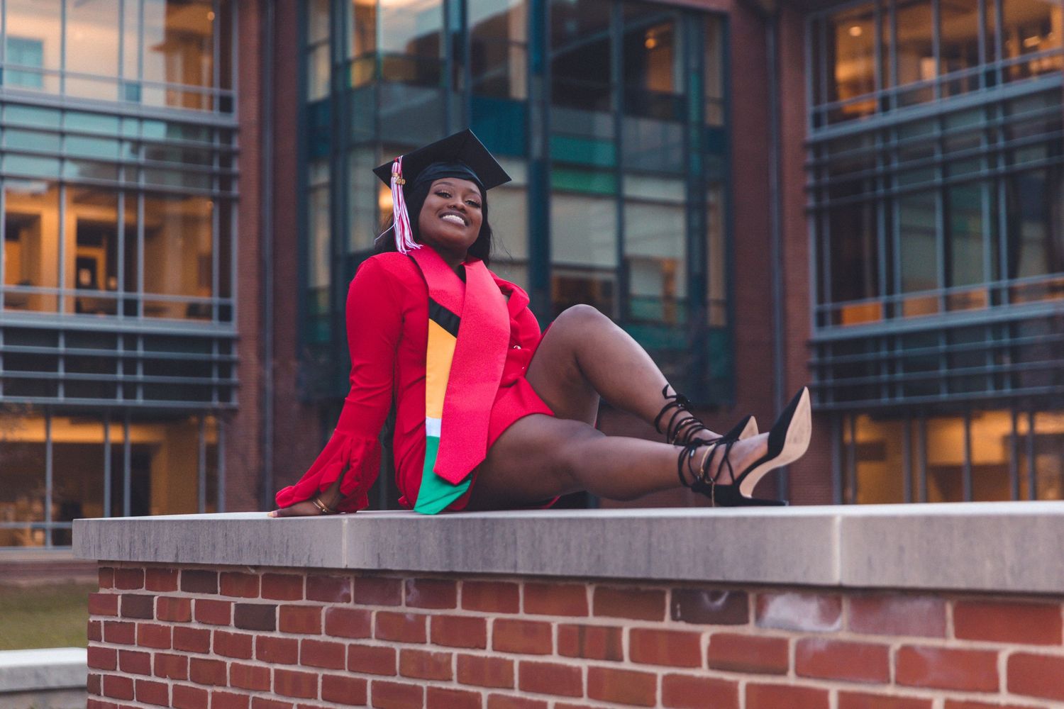 Smiling graduate in red gown and cap sitting on brick wall outside modern building.