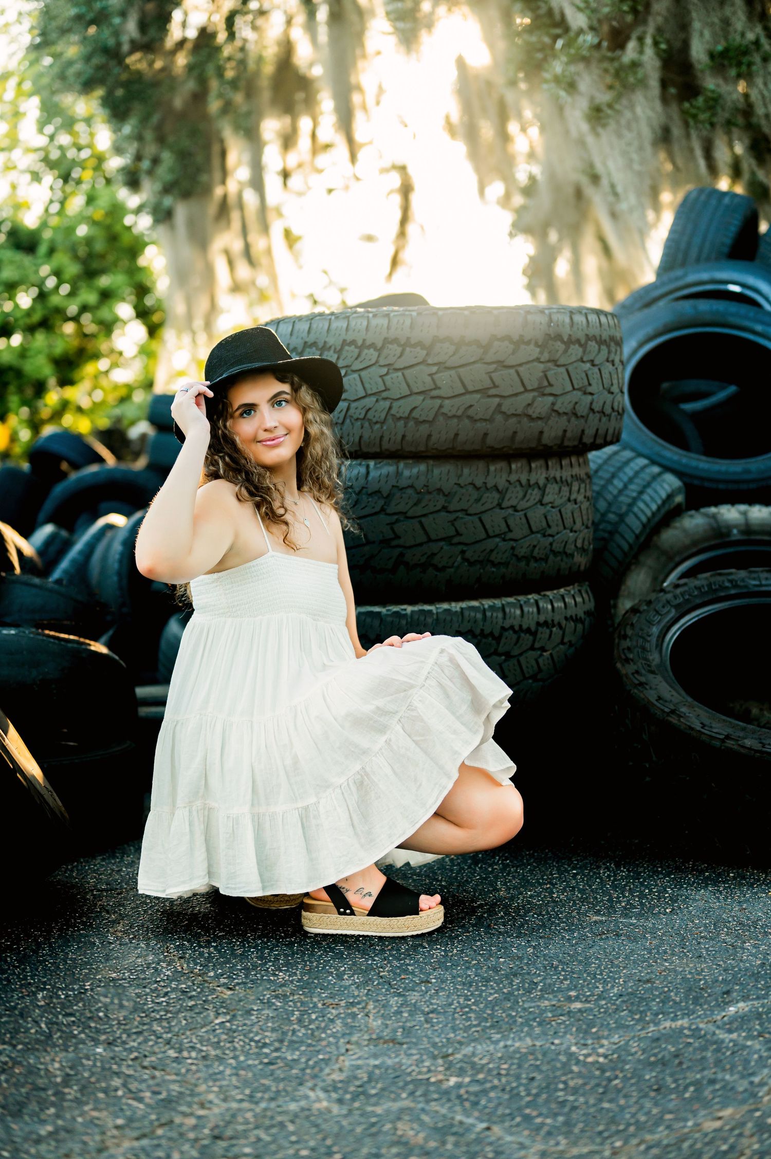 Woman in hat and white dress poses casually against stacked tires.