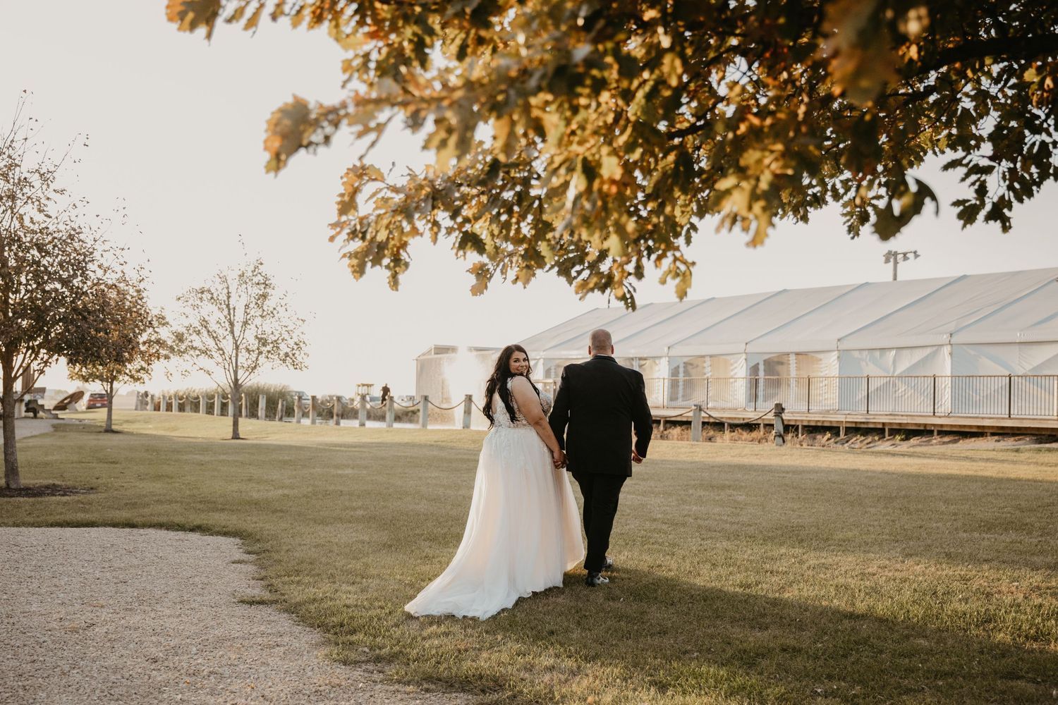 bride and groom walk away from photographer, she looks back and smiles as they walk under tree back towards the venue to join their party