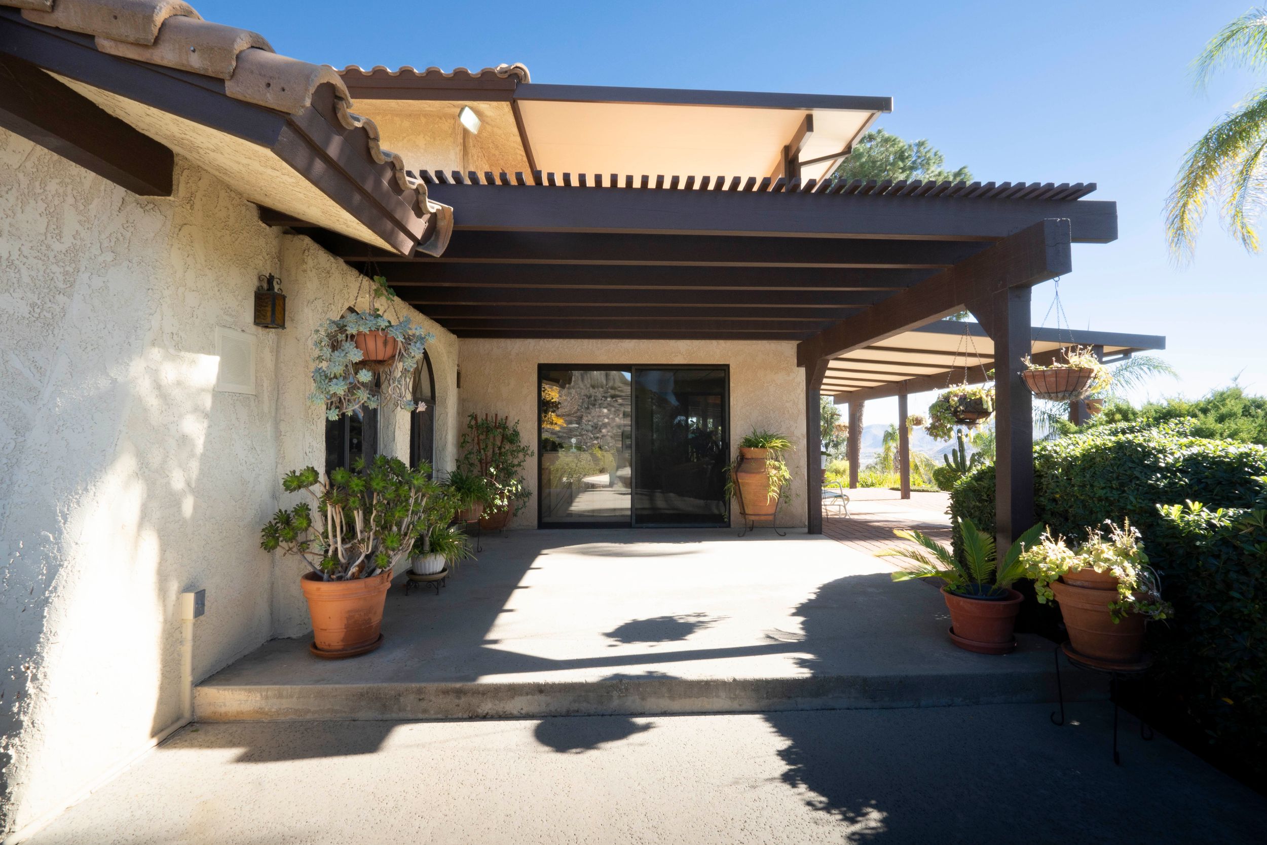 Spanish-style home entrance with a wooden pergola, terracotta pots, and lush landscaping.