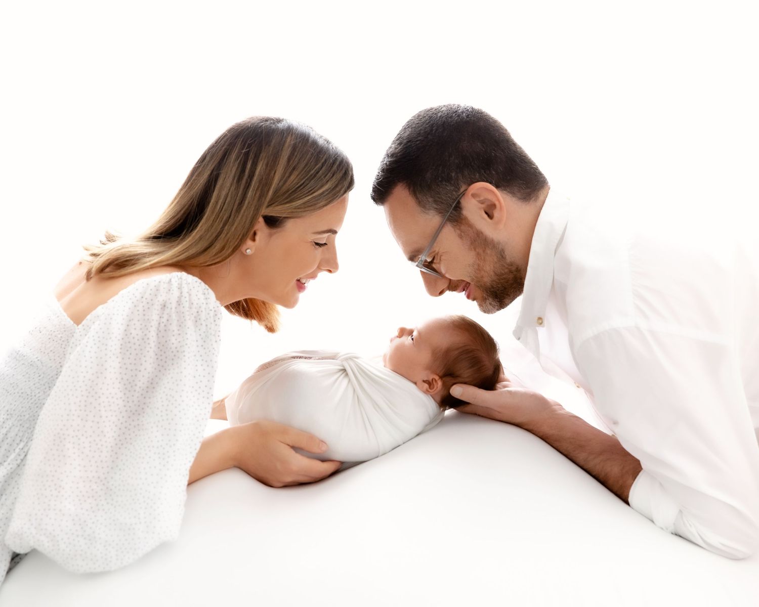 Parents in white clothing lean in close to admire their newborn baby.
