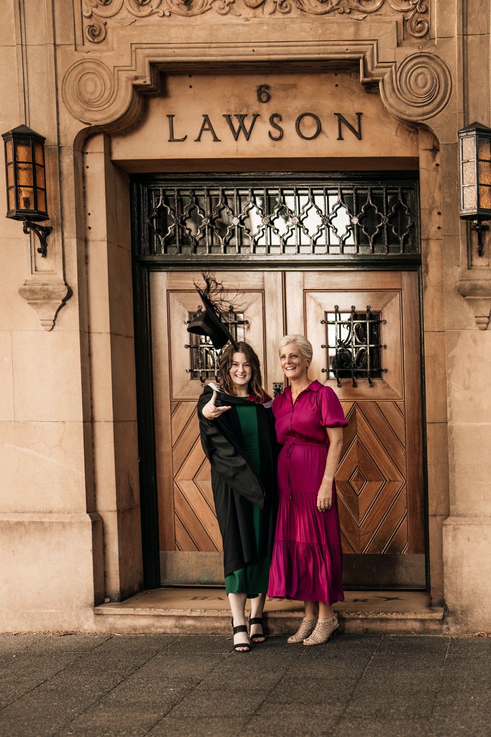 Two people pose elegantly in front of an ornate doorway marked 'Lawson' wearing formal green and pink dresses.