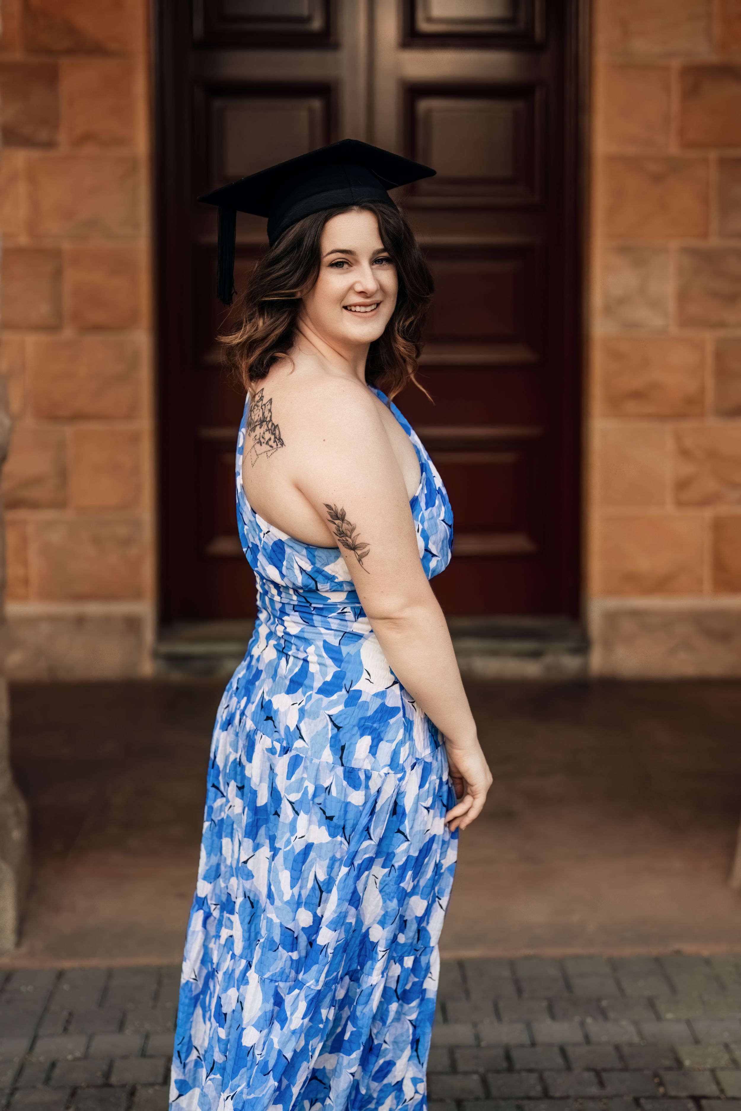 Graduate in blue floral dress and cap poses confidently showing tattoo near stone building entrance.