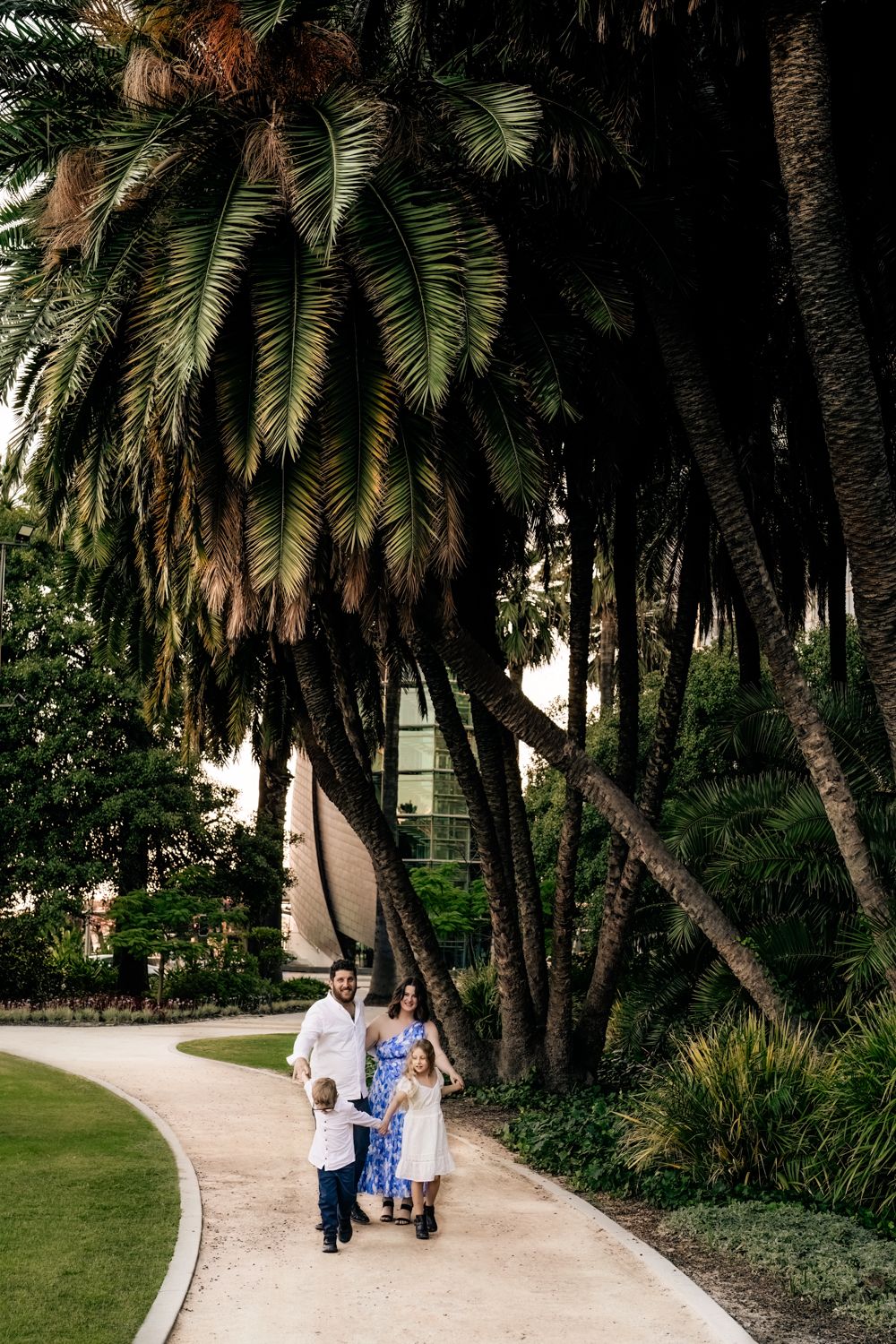 People walking along a curved path beneath tall palm trees in a tropical garden setting.