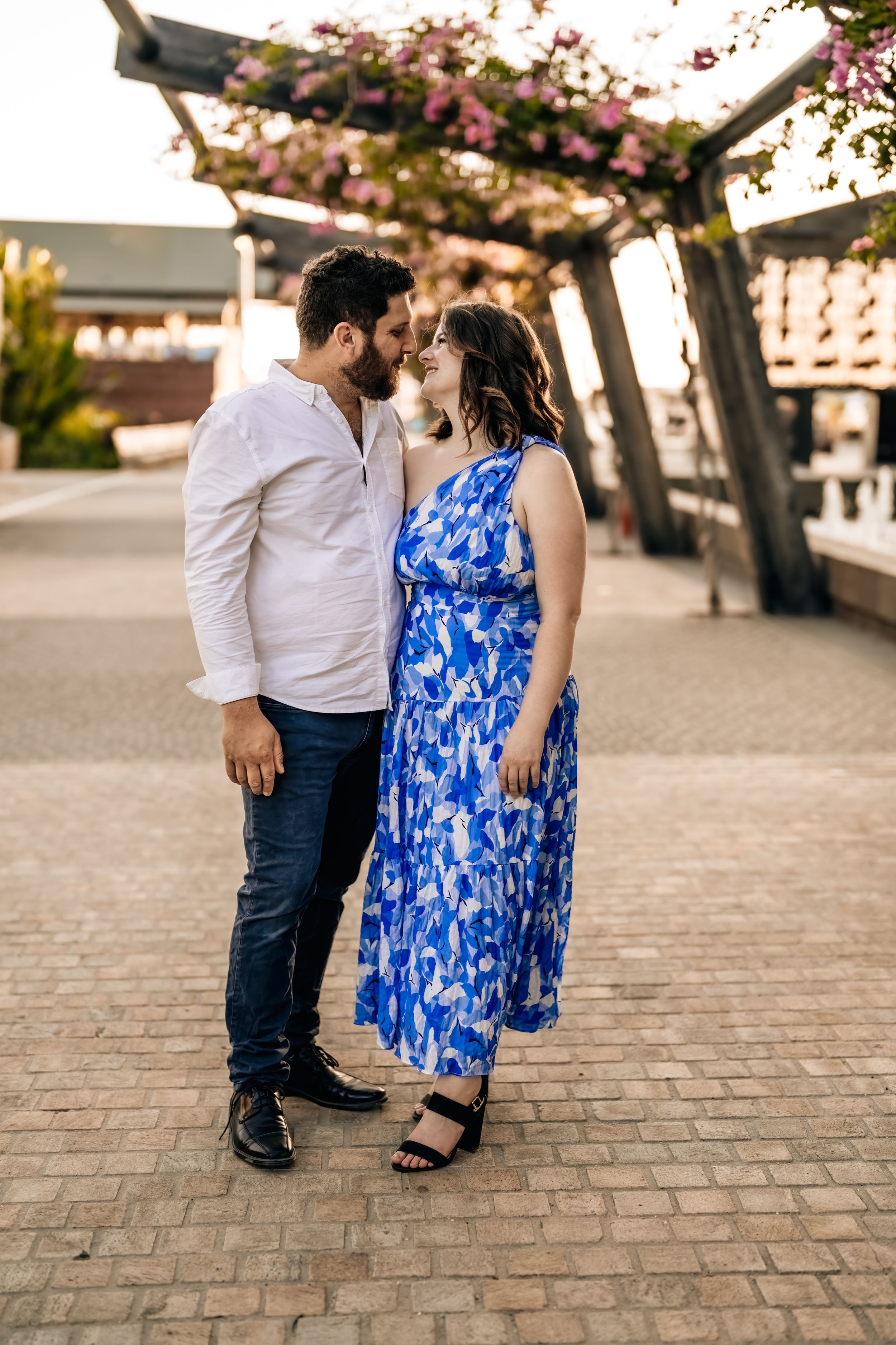 Couple poses together on cobblestone pavement with blooming pink trees overhead.