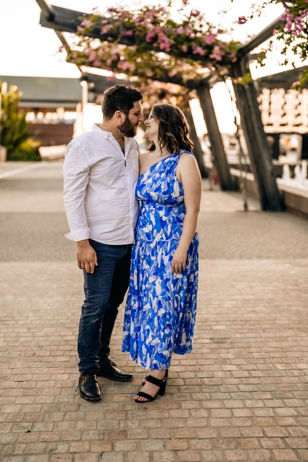Couple poses together on cobblestone pavement with blooming pink trees overhead.