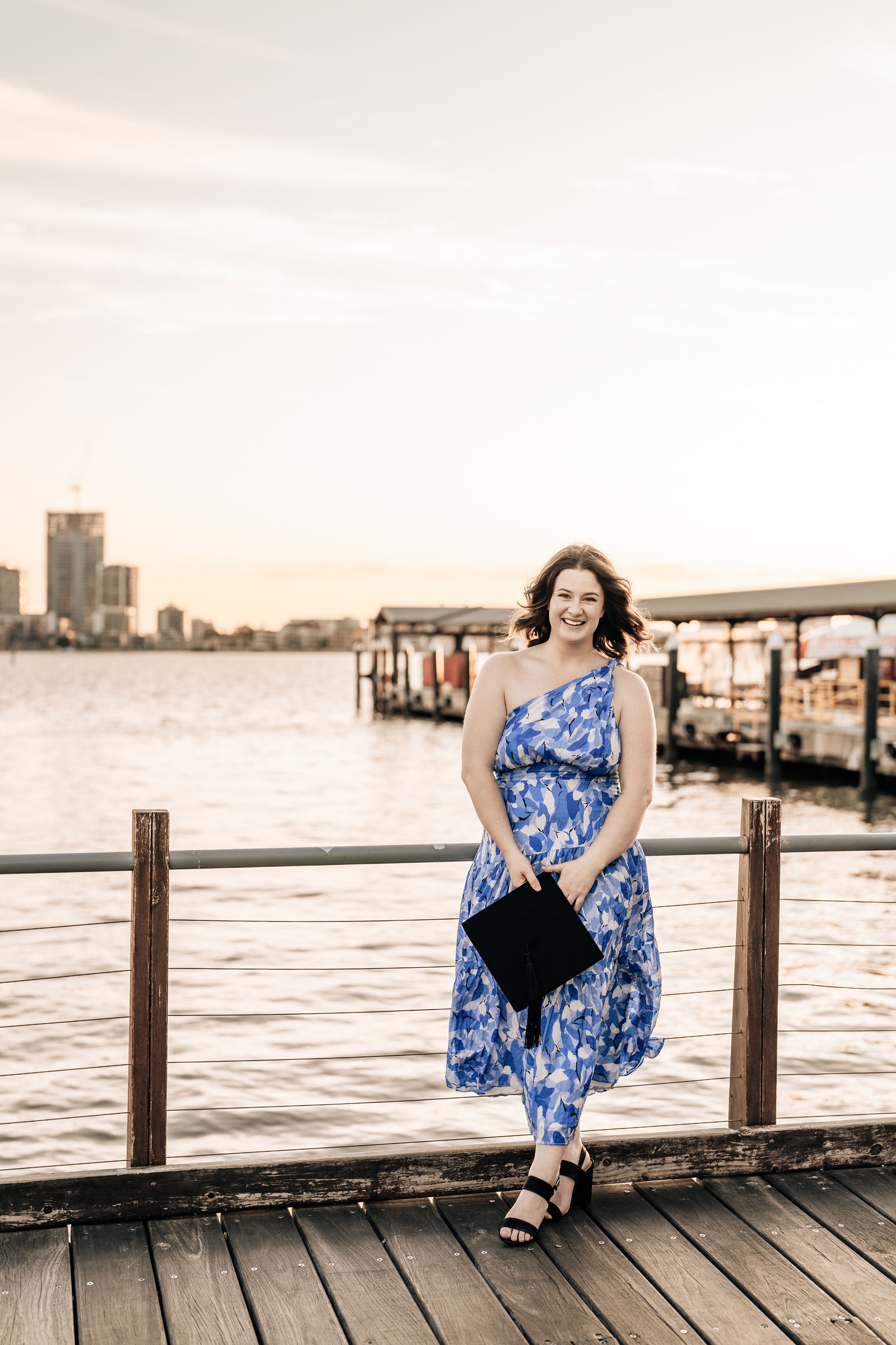 Woman in blue floral dress sitting casually on wooden pier at sunset.