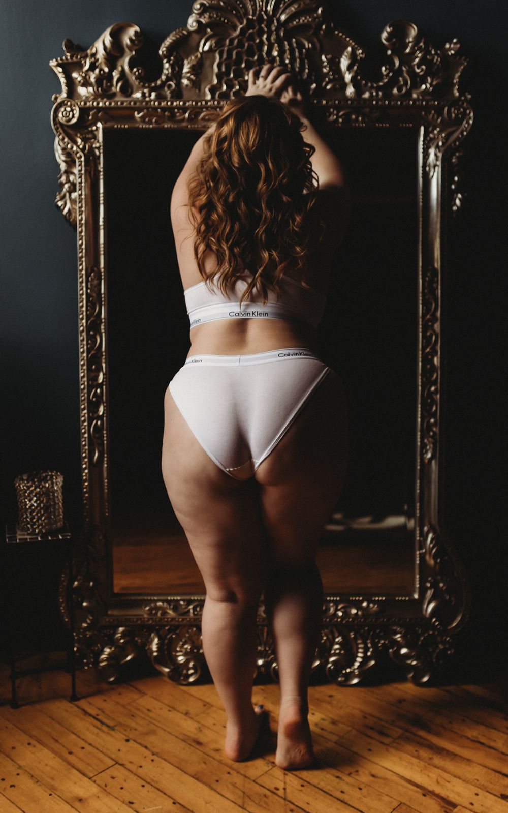 Backlit figure in white lingerie stands before an ornate gold baroque mirror on wooden floor.