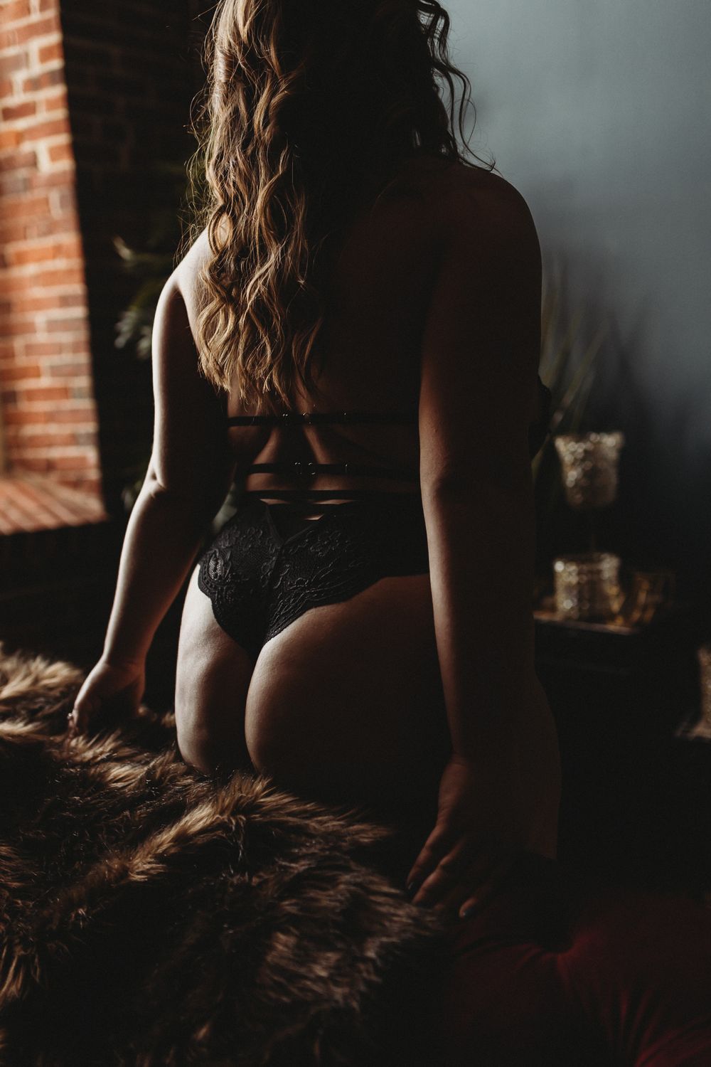 Silhouette of a person in black lingerie kneeling on a fur rug near a brick wall in moody lighting.