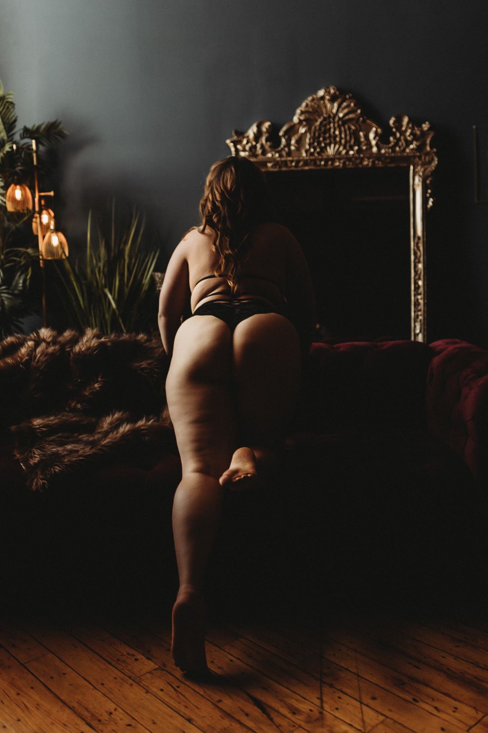 Artistic boudoir photo with ornate gold mirror and moody lighting on wooden floor.
