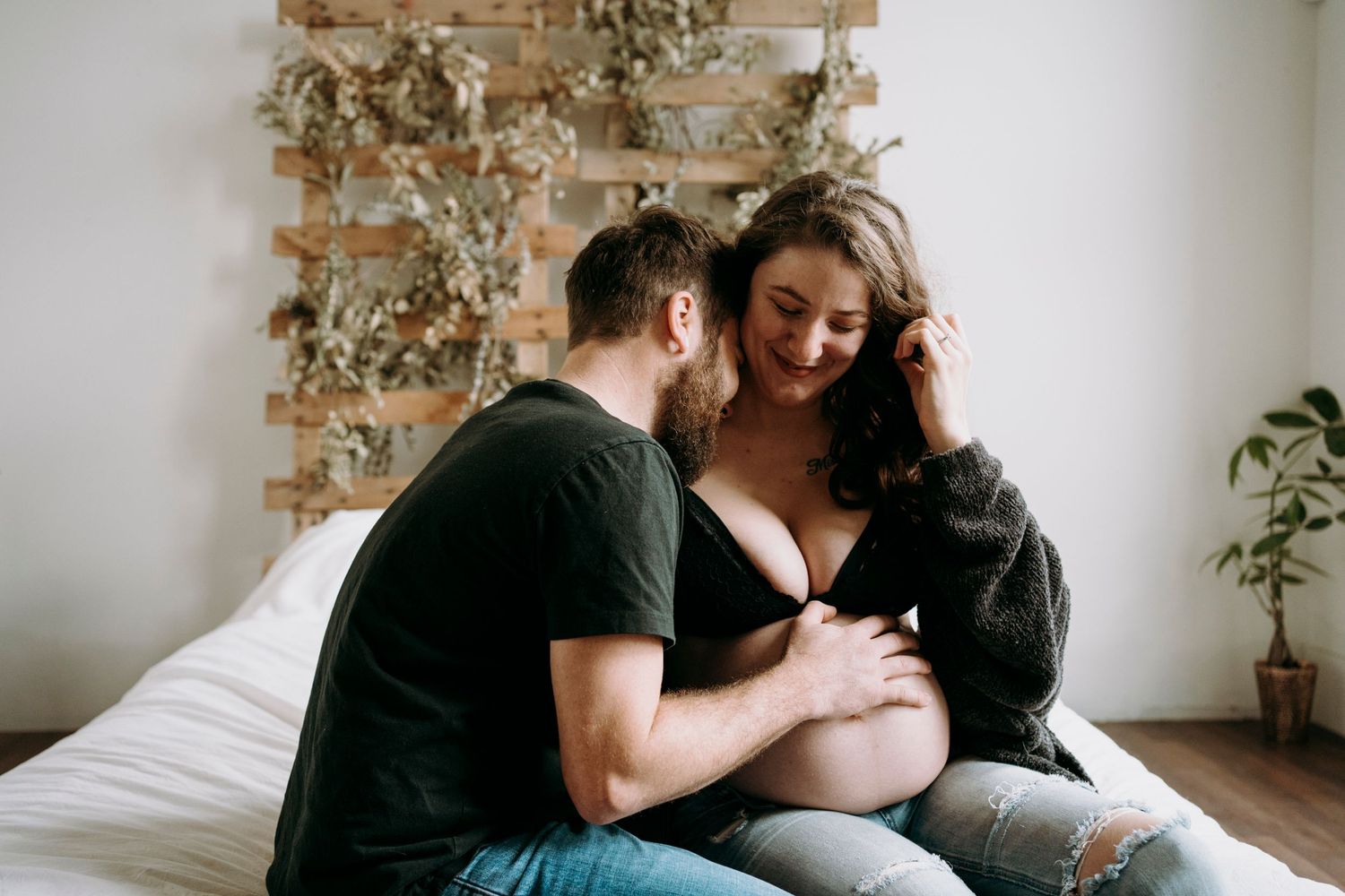 Intimate maternity photo of couple sitting on bed with hanging plants in background.