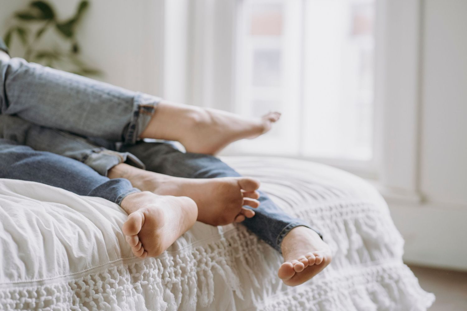 Bare feet resting on white bedding near window with natural light.