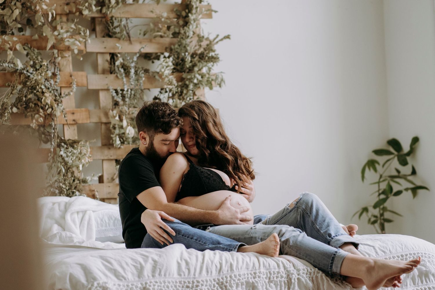 Intimate couple cuddles on white bed with decorative wooden wall panel and greenery in background.