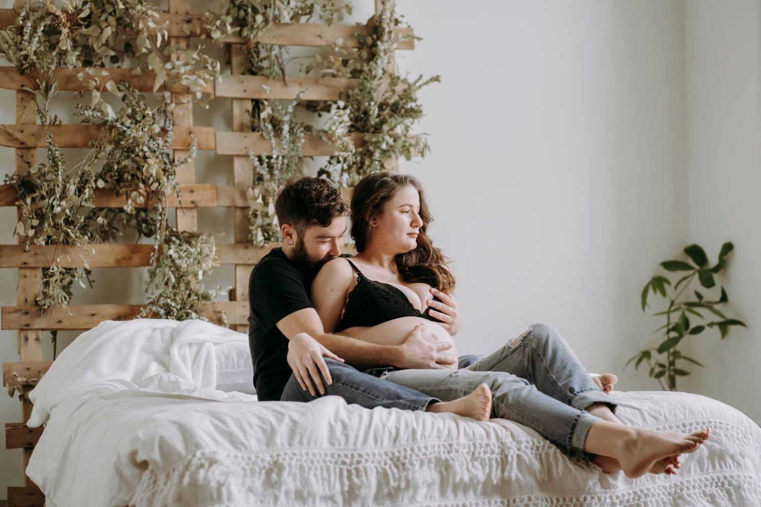 Couple lounging together on white bedding against rustic wooden wall decor with plants.