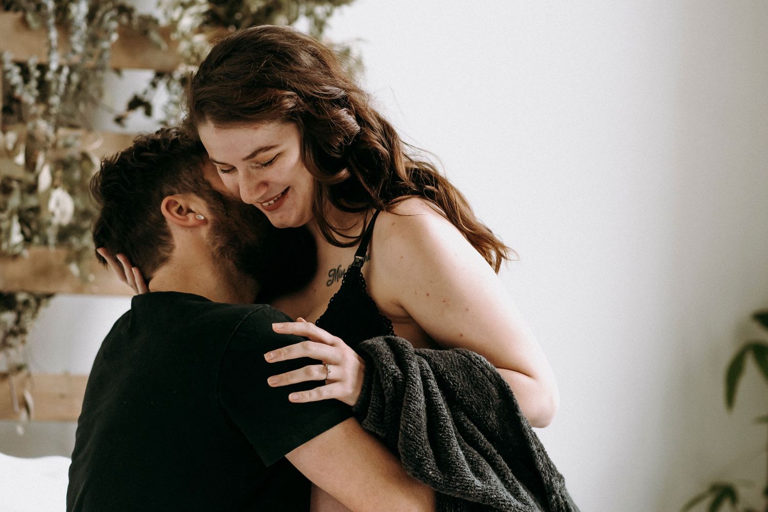Intimate couple sharing tender moment on white bed with wooden wall decor and plants.