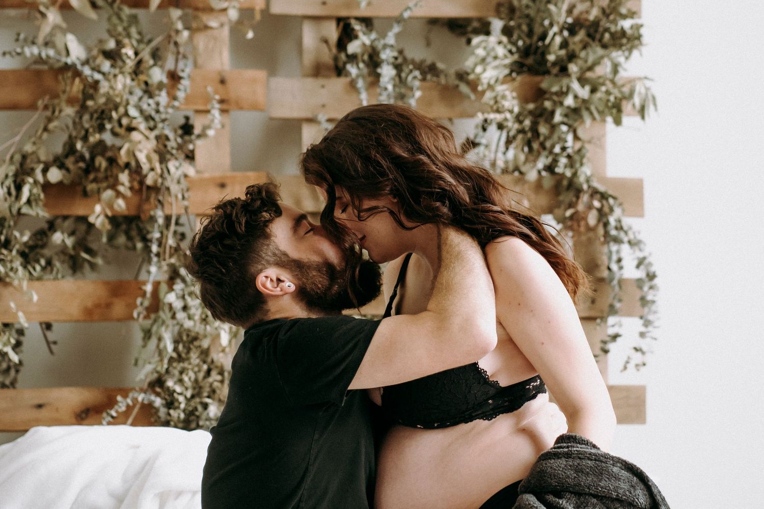 Couple sharing intimate moment on white bedding with rustic wood wall decor and plants.