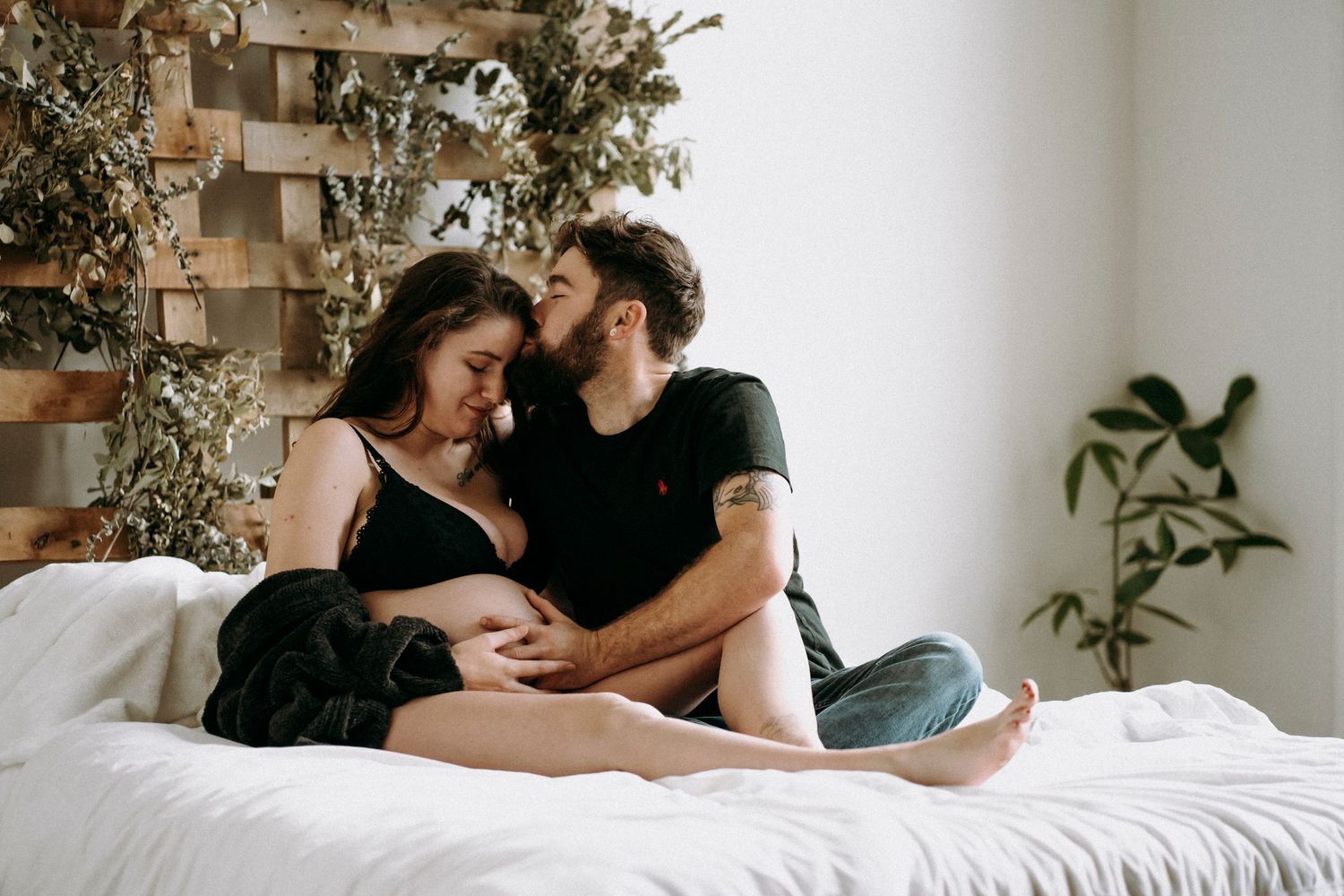 Couple relaxing together on white bed against wooden wall panel with hanging plants.