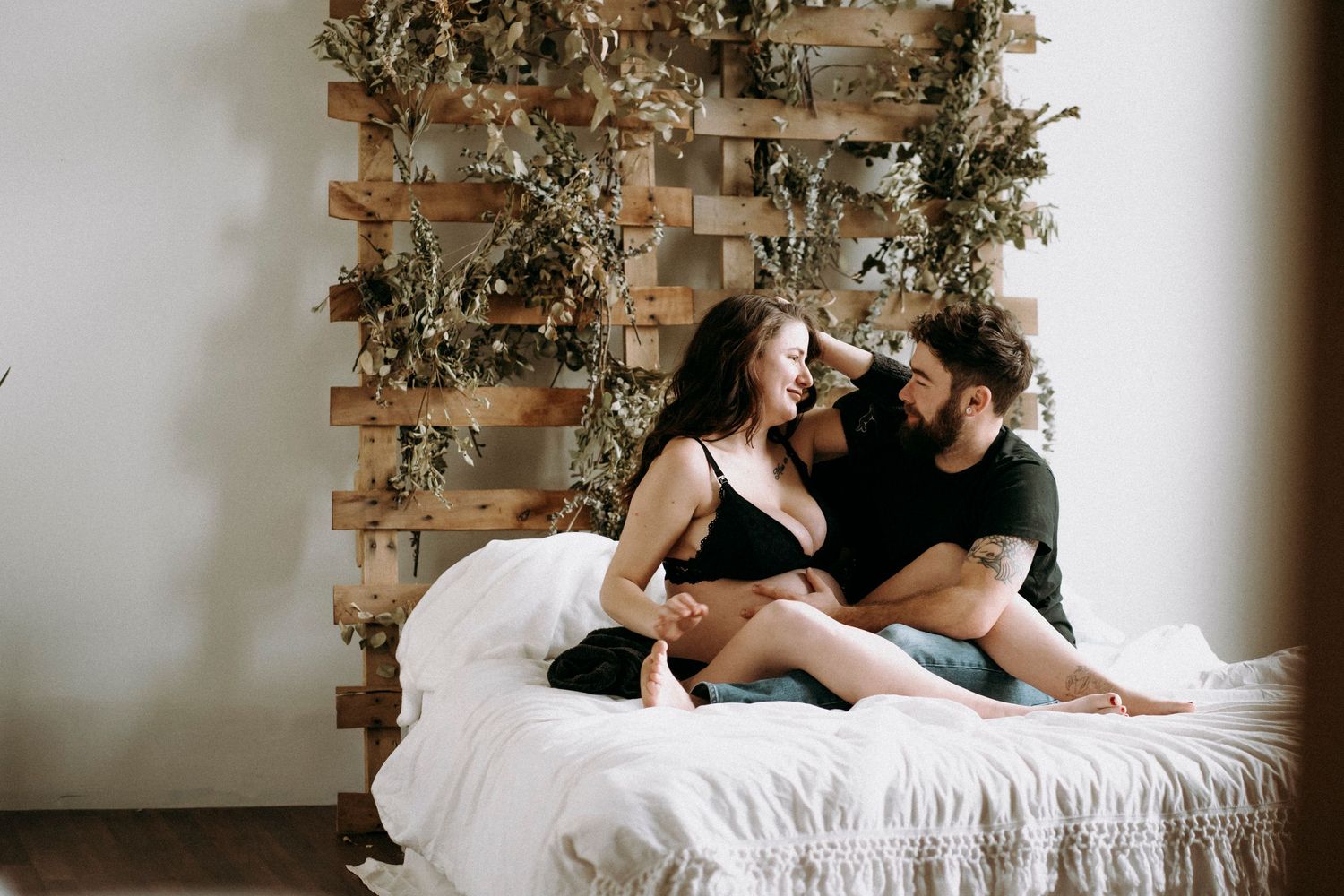 Couple sitting together on white bed with wooden wall decor and plants in background.