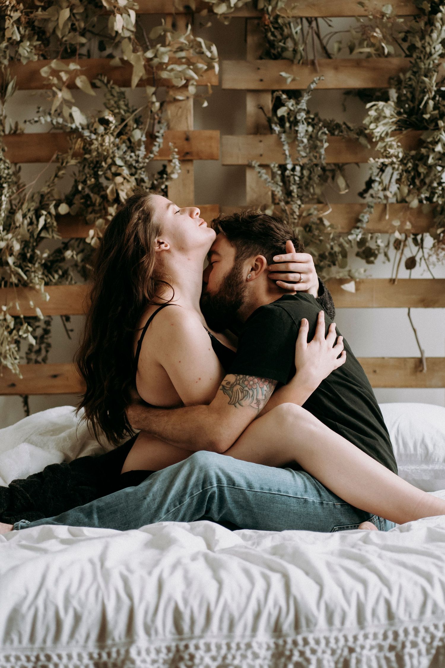 Romantic couple embracing on a bed with dried floral wall decor in the background.