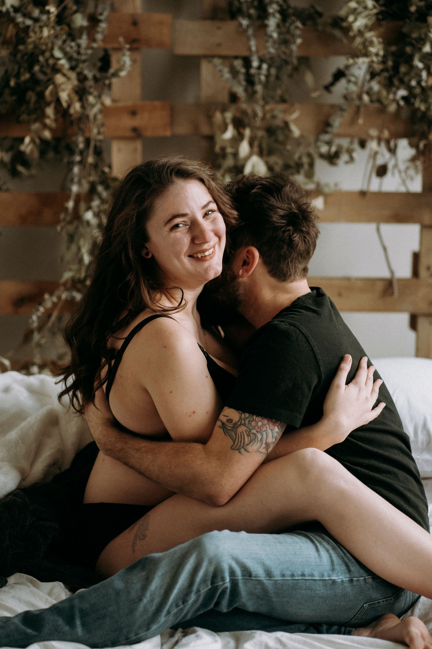 Couple sharing intimate moment on bed with rustic wooden wall backdrop decorated with dried flowers.